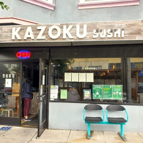 Kazoku Sushi storefront with an "OPEN" sign, a person inside, and a two-seat bench outside.