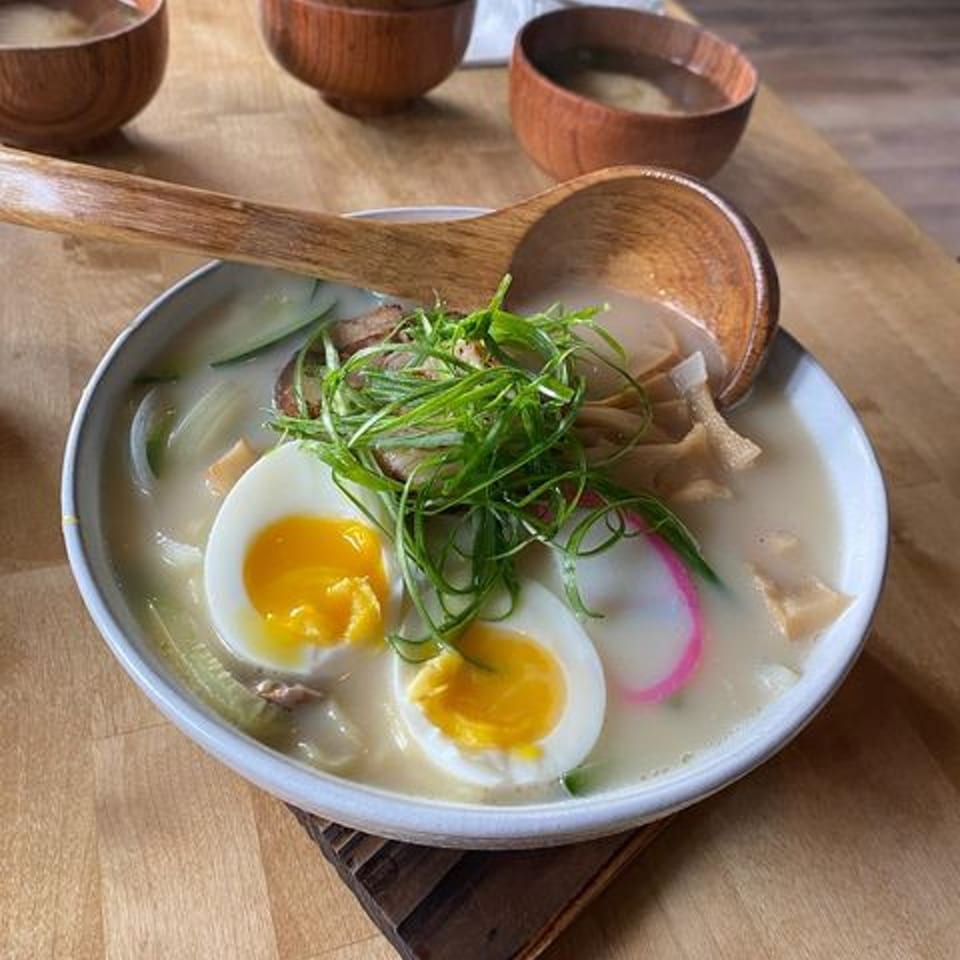 A bowl of ramen with sliced eggs, green onions, and fish cake, served with a wooden spoon.