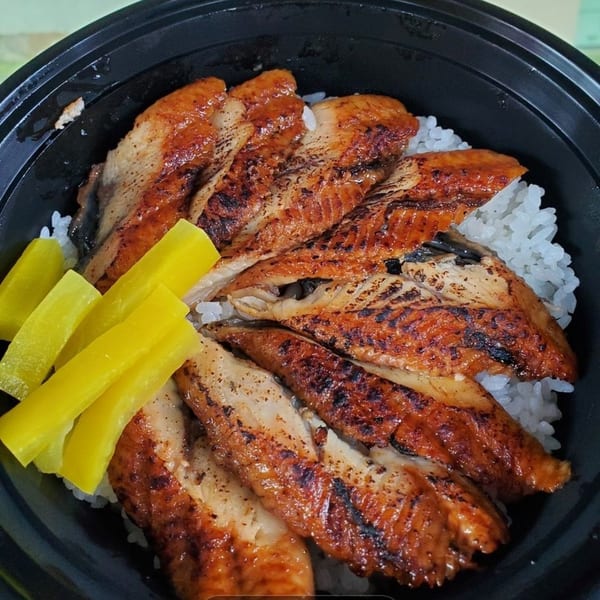 Grilled eel (unagi) over white rice with yellow pickled daikon in a black bowl.