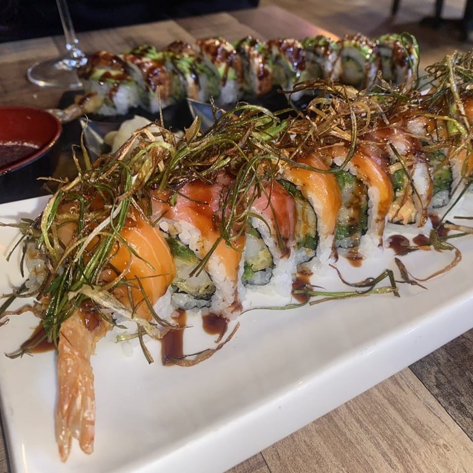 Two long plates of sushi rolls, one topped with salmon and crispy fried greens, the other with avocado.